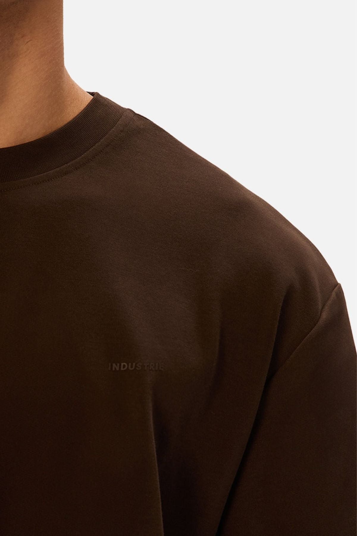 Close-up of a person wearing the Industrie The Newport Tee in dark brown. The oversized, heavy-weight fit is shown from shoulder to chest, with "INDUSTRIE" subtly visible against a plain background.
