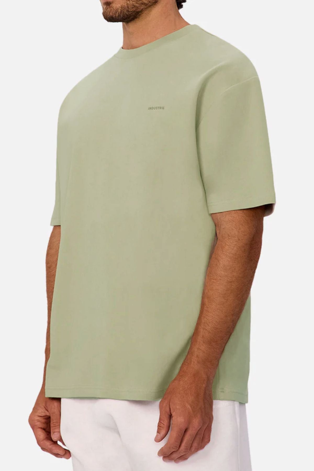 A man in an oversized Industrie The Newport Tee in light green—heavyweight, 100% cotton, with minimal chest text—and white pants stands against a plain light background.