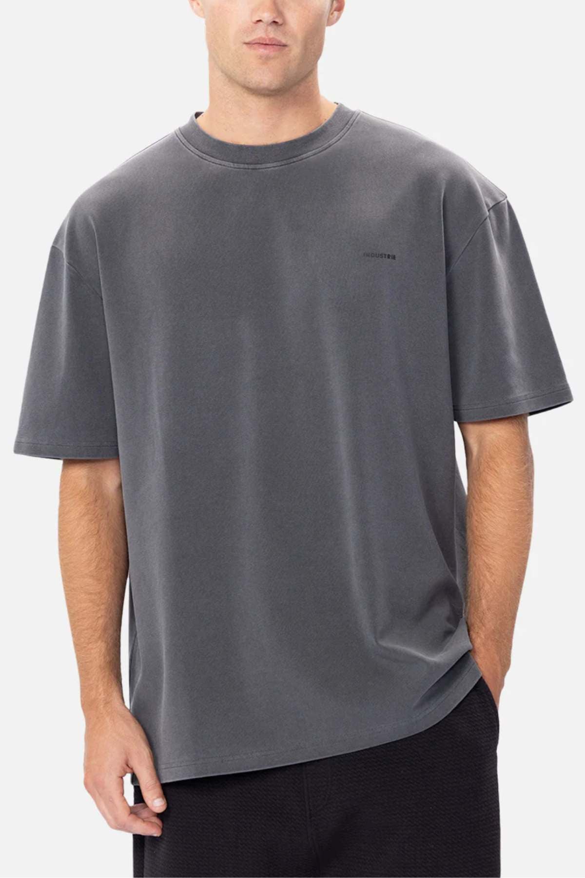 Wearing Industrie The Newport Tee in oversized dark gray with a subtle text design on the left chest, made from 100% cotton, the person stands against a plain white background in black pants.