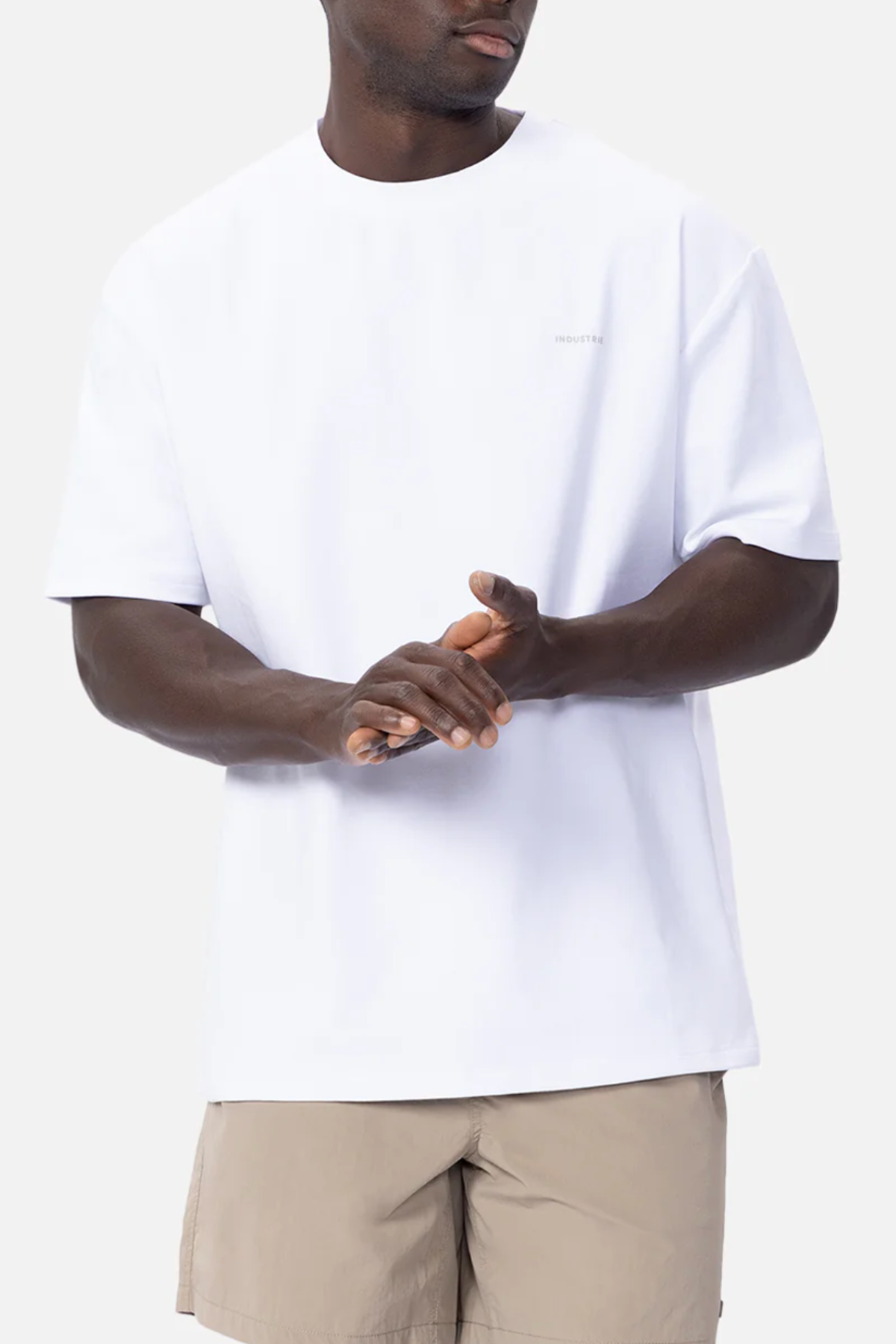 A person wearing an Industrie The Newport Tee in plain white, made of 100% cotton fabric, and light beige shorts is standing against a white background. The individual's face is not visible, and they are clasping their hands together in front of them.