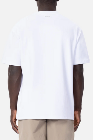 A person with short hair is standing with their back to the camera, showcasing an oversized fit Industrie The Newport Tee in plain white and light beige pants. The background is a clean and minimalist white.