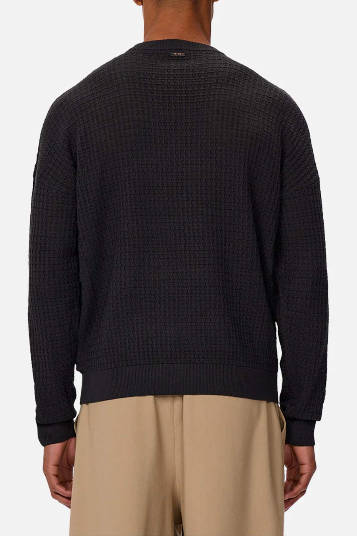 A person is shown from the back against a plain light background wearing the Industrie The Nunez Long Sleeve Knit, a textured black 100% cotton sweater, paired with beige pants.