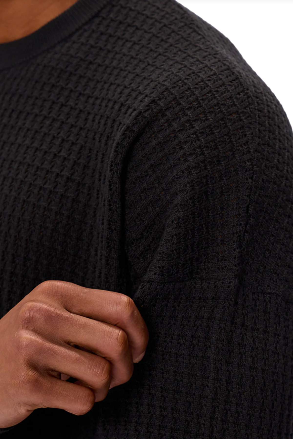 Close-up of a person wearing the Industrie The Nunez Long Sleeve Knit in black, featuring a textured 100% cotton waffle-knit fabric. One hand holds the sleeve, showcasing the detailed pattern and versatile style of this shirt.