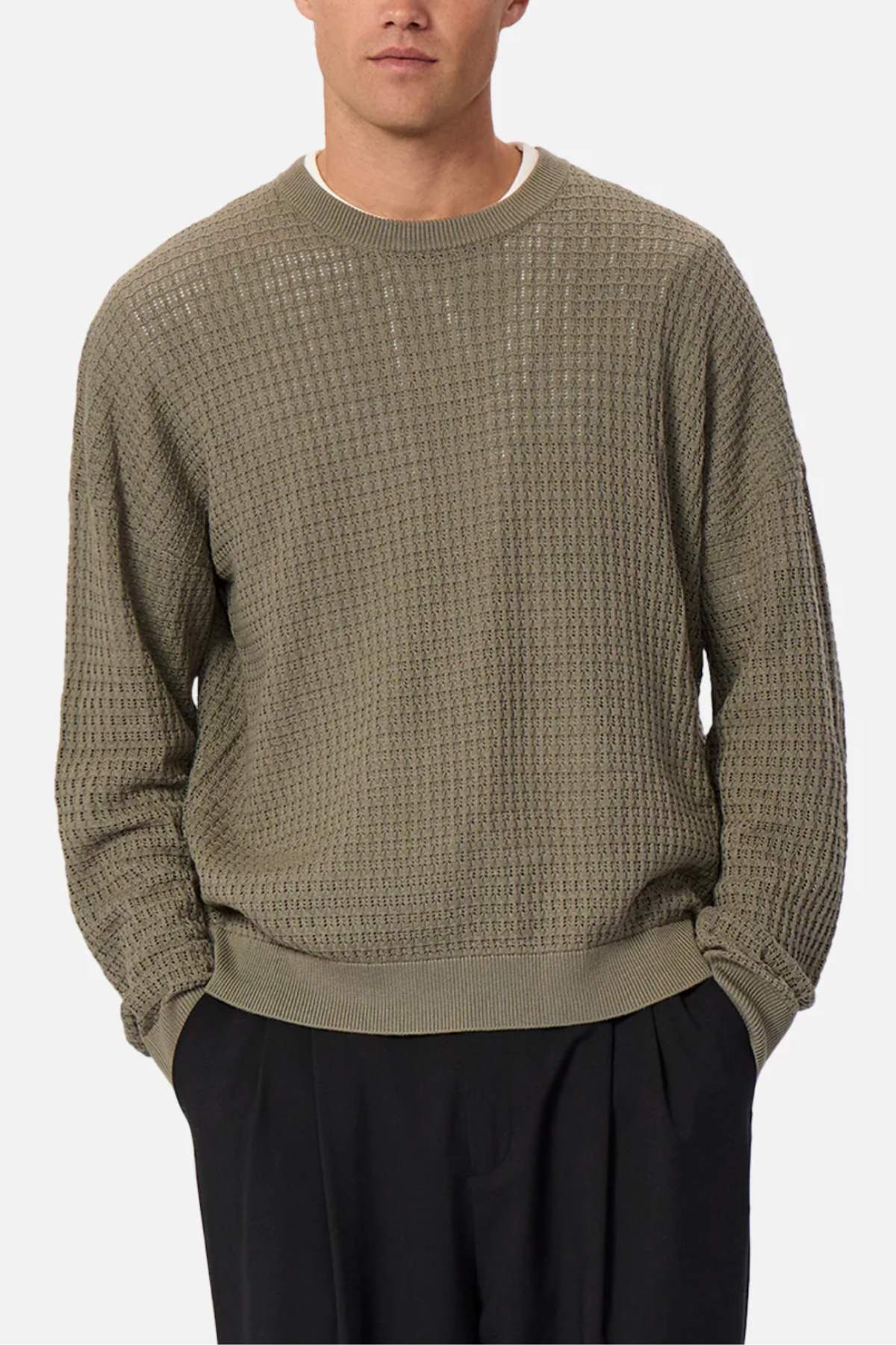 A person wears the Industrie The Nunez Long Sleeve Knit, a green textured sweater with ribbed cuffs and a crew neck, made from 100% cotton. It pairs effortlessly with black pants against a plain off-white backdrop, enhancing its relaxed charm as hands rest in pockets.