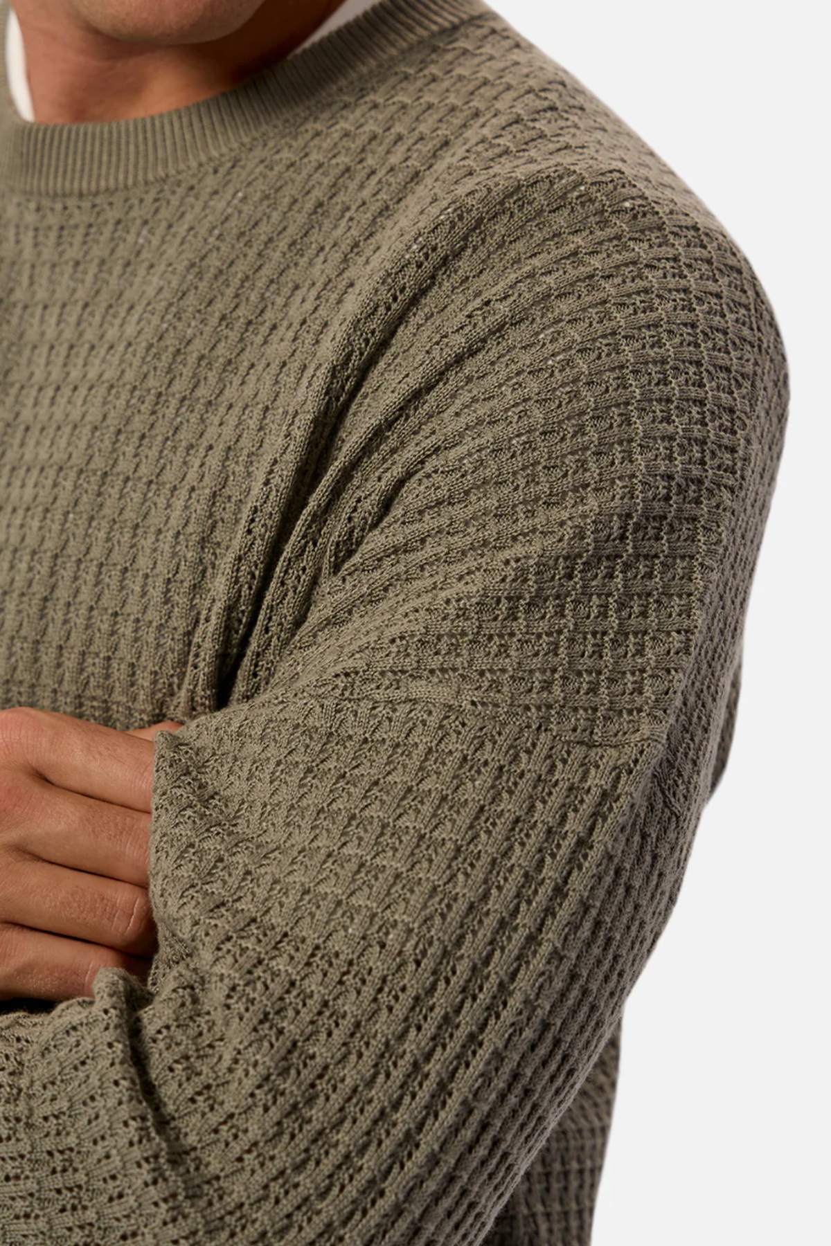 A person wears the Industrie The Nunez Long Sleeve Knit sweater in textured olive green, layered over a white shirt. Their arms are crossed, showcasing the grid-like pattern in 100% cotton with ribbed cuffs against a neutral background.