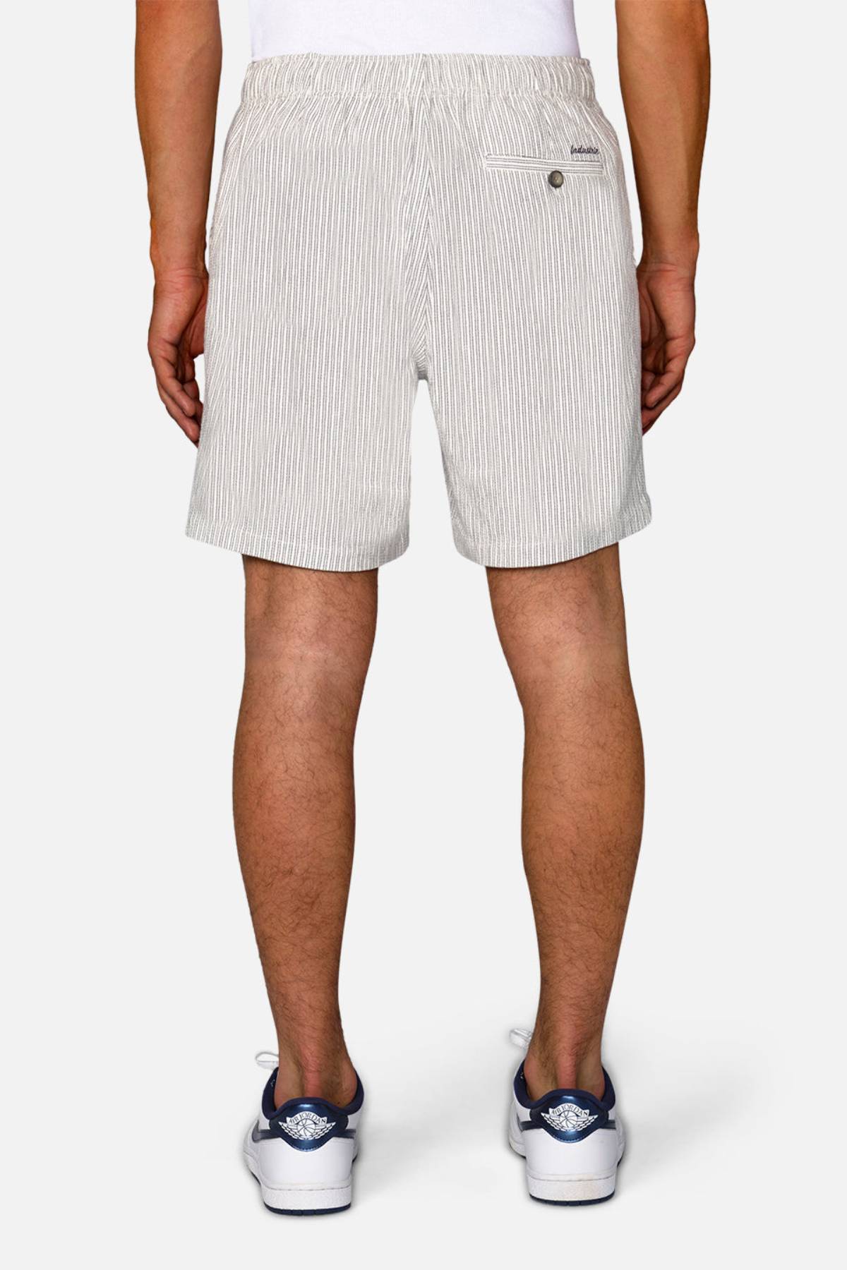 A person faces away from the camera, wearing Industrie The Ortenza Short in light gray with an elastic waistband, textured fabric, and a single buttoned back pocket, paired with a white shirt and white sneakers with blue accents.