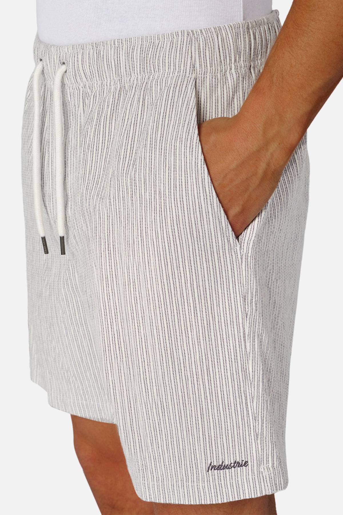 A person wears light-colored, vertically striped Industrie The Ortenza Short with a hand in the pocket, featuring a subtle embroidered logo near the hem, paired with a crisp white shirt.