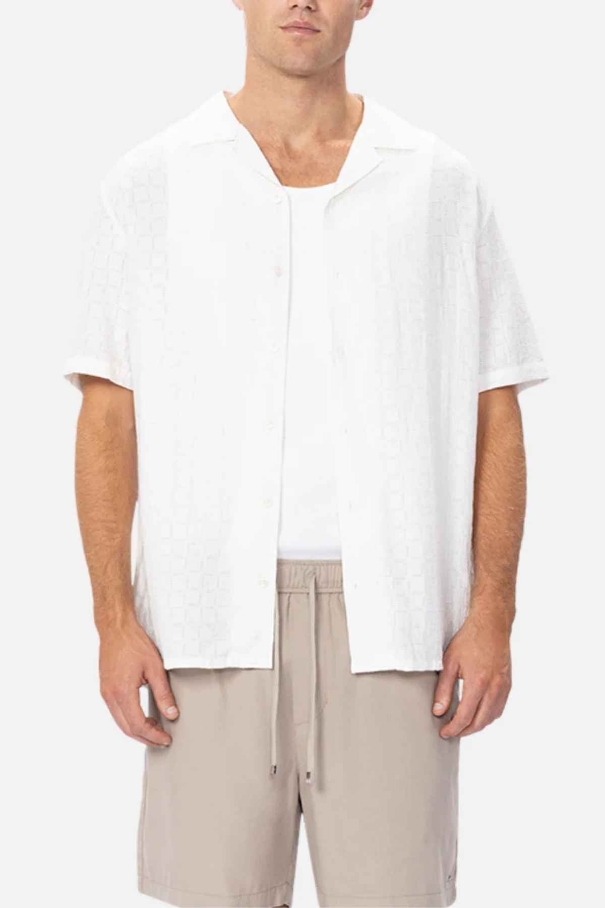 A person wearing a breathable Industrie The Ortigia Short Sleeve Shirt, a white short-sleeved button-up over a tank top, is paired with beige shorts, standing against a plain background.