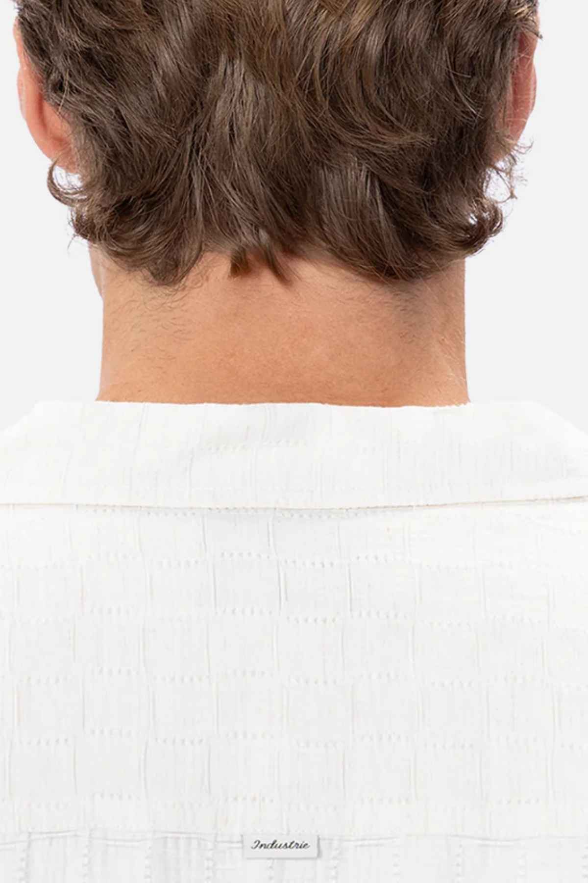 The image showcases the back of a person's head and shoulders, featuring wavy brown hair draping over an Industrie The Ortigia Short Sleeve Shirt. This white, textured party shirt is collared and crafted from breathable fabric, all presented against a plain white backdrop.