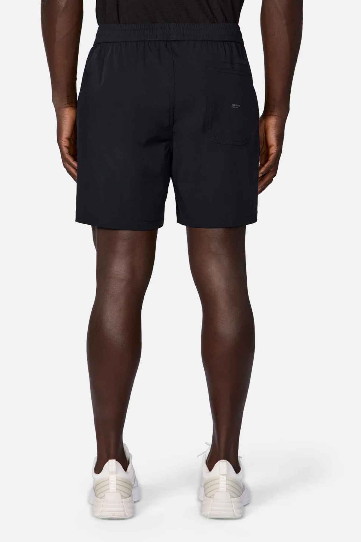 A person wearing the Industrie The Owens Short in a stylish black design, featuring an elasticised waistband and a back pocket on the right side, stands facing away while pairing them with white sneakers. The quick-drying fabric of the shorts stands out against the plain white background.
