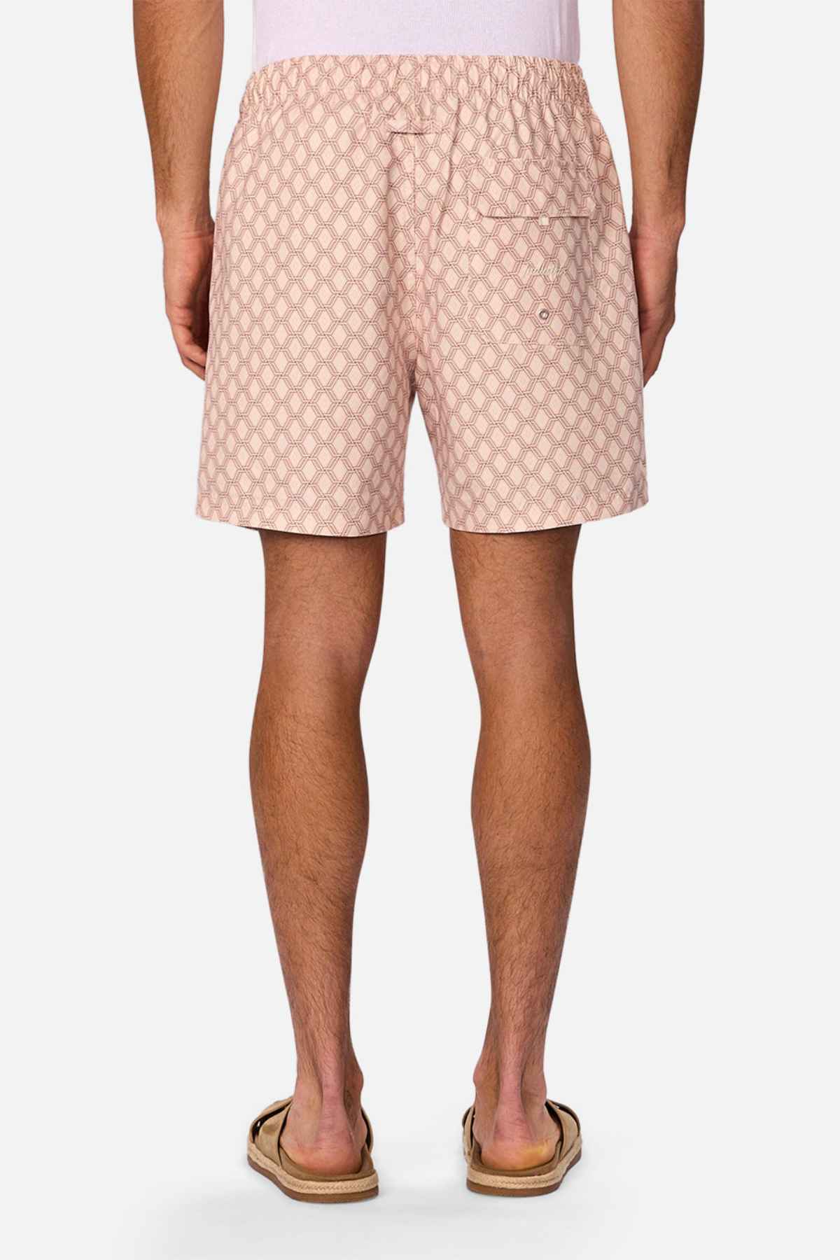A person, only visible from the waist down, is wearing Industrie The Panama Swim Short in light pink with a diamond pattern and flip-flops. The men's swimwear features an elastic waistband and a buttoned back pocket.