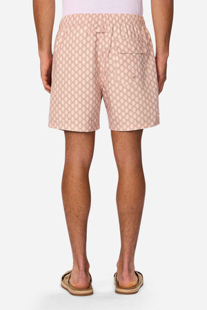 A person, only visible from the waist down, is wearing Industrie The Panama Swim Short in light pink with a diamond pattern and flip-flops. The men's swimwear features an elastic waistband and a buttoned back pocket.