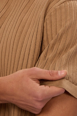 A close-up of a person holding the sleeve of the Industrie The Poolside Polo in light brown ribbed cotton knit, ideal for casual wear. The textured fabric and pattern are highlighted, showing only a hand, arm, and part of the upper body.