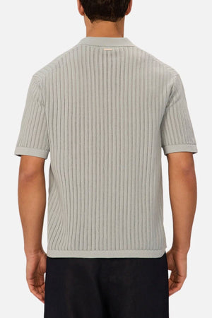 A man is seen from behind wearing Industrie The Poolside Polo in light gray, a short-sleeved cotton knit with subtle vertical stripes and a collar, paired with dark pants—ideal for casual occasions.