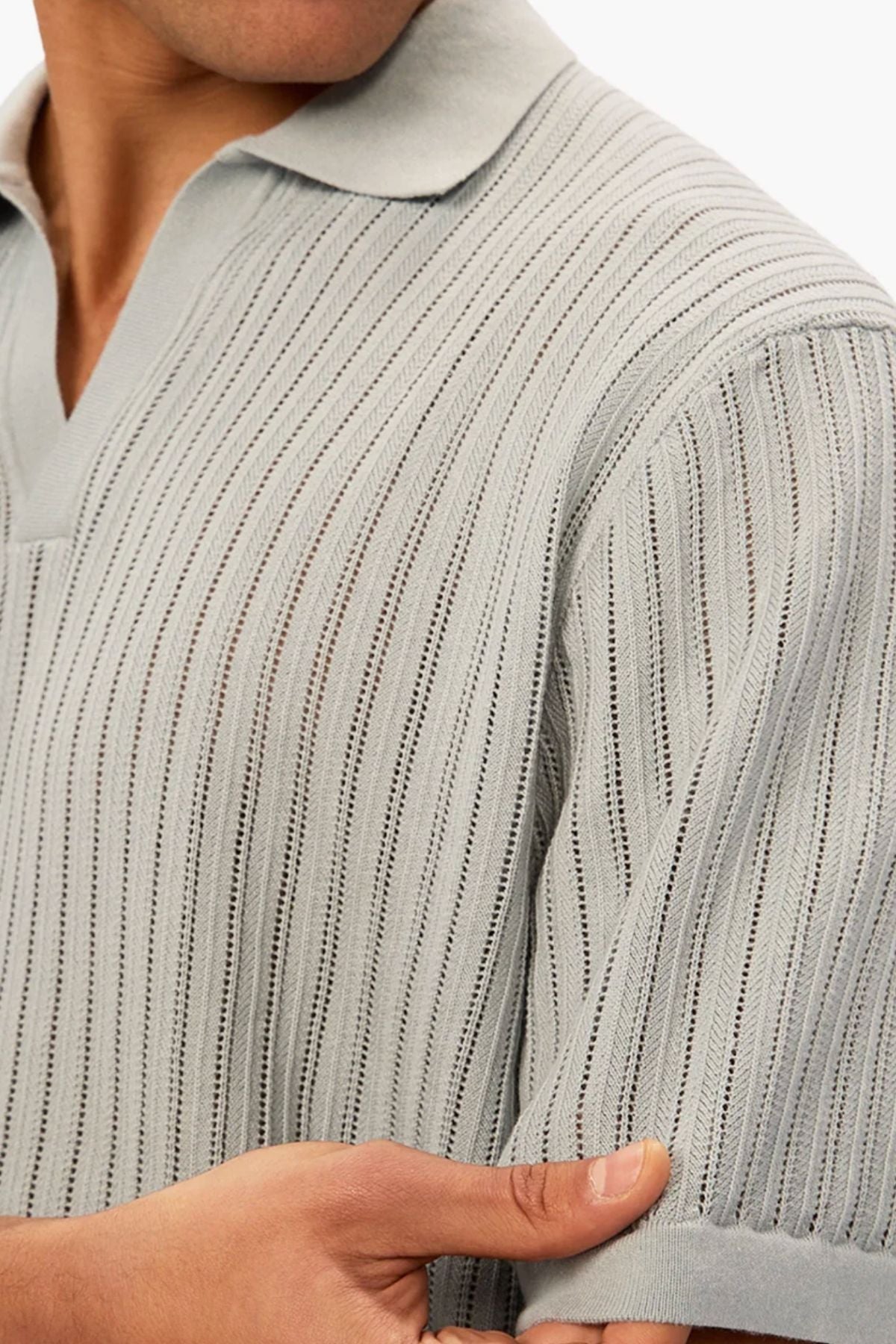 A person wears the Industrie The Poolside Polo—a light grey, short-sleeve, cotton knit with a subtle vertical pattern. They hold the sleeve's edge with one hand, and their face is partially out of frame.