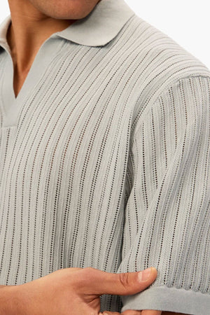 A person wears the Industrie The Poolside Polo—a light grey, short-sleeve, cotton knit with a subtle vertical pattern. They hold the sleeve's edge with one hand, and their face is partially out of frame.