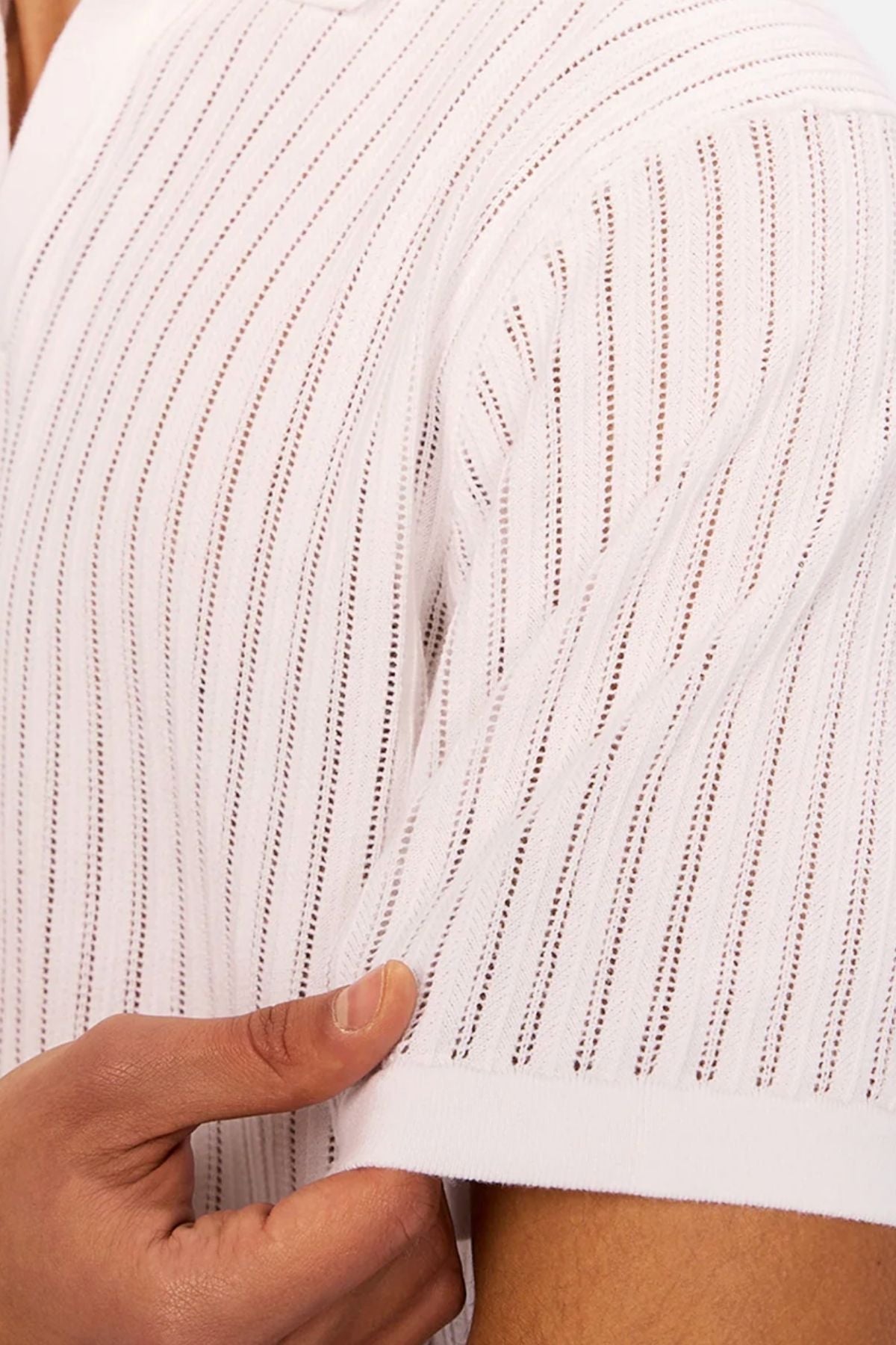 A close-up of a person wearing Industrie The Poolside Polo—a white, short-sleeved, cotton-knit top with vertical perforated stripes—gently holding the sleeve edge to highlight its effortless casual style.