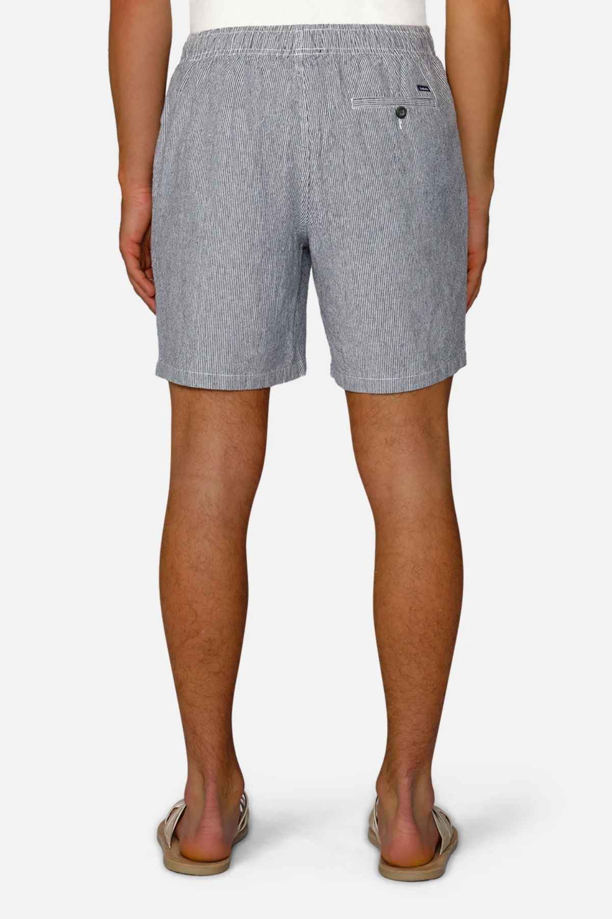 Rear view of a person wearing Industrie The Portico Linen Short, featuring a subtle pale blue stripe design. These shorts are paired with a crisp white T-shirt and flip-flops as they stand against a clean white background, perfectly capturing the essence of a summer wardrobe.