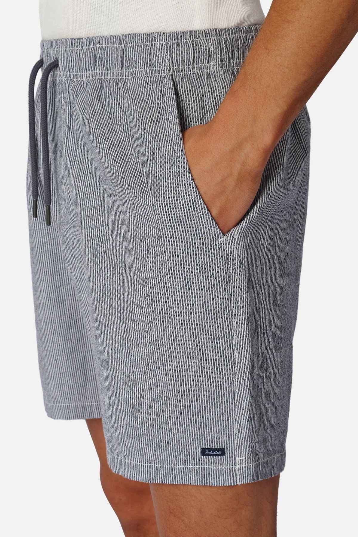 A person wearing Industrie The Portico Linen Short featuring a pale blue stripe design stands with hands in pockets against a plain background—a must-have for any summer wardrobe.