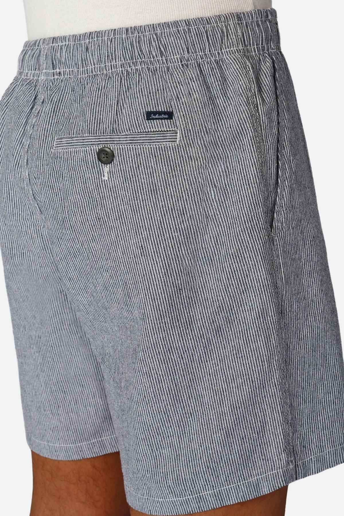 A person is wearing Industrie The Portico Linen Short featuring a pale blue stripe pattern and an elastic waistband. Ideal for a summer wardrobe, these shorts boast a back pocket with a button and are accented with a small logo tag above it. The fabric looks lightweight and comfortable.