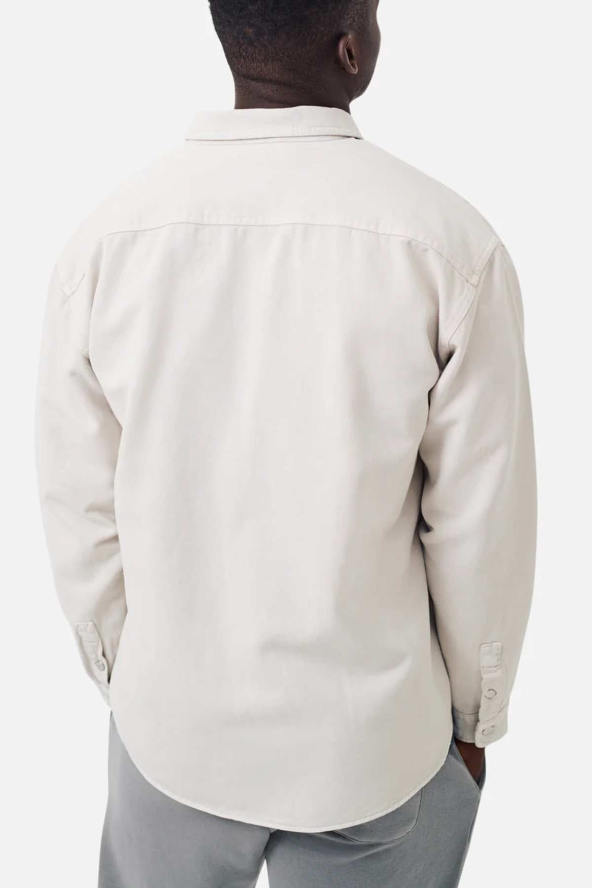 The image displays a person from the back wearing Industrie The Portland Shirt, a light beige cotton-linen overshirt with a relaxed fit, buttoned cuffs, and classic collar. They pair it with light gray pants against a plain white background.
