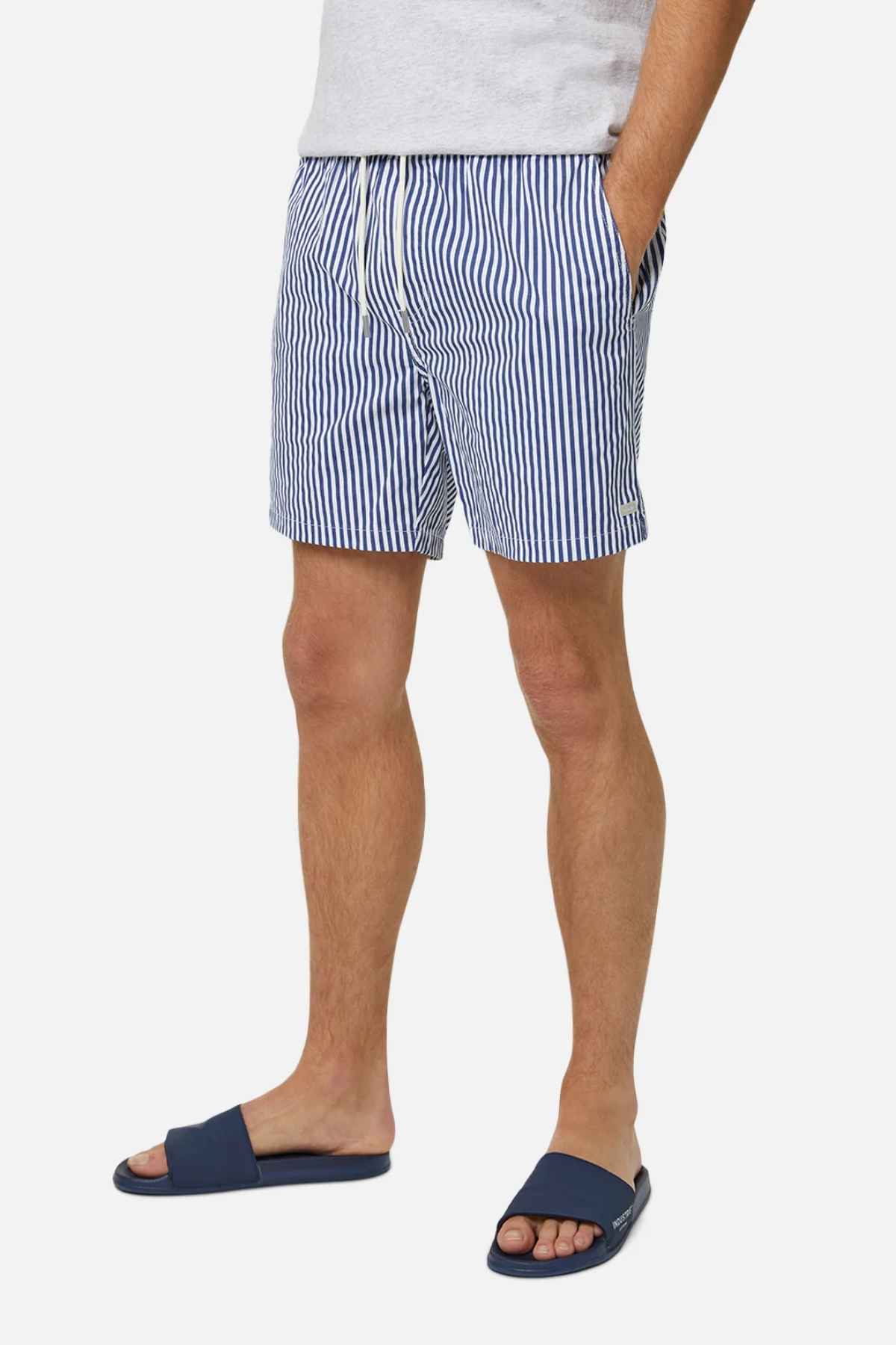 A person wearing Industrie The Princeton Bahama Short in a blue and white striped pattern pairs it with a light gray t-shirt made from a comfortable fabric blend, standing against a plain white background. With their hands in the pockets and sporting blue slide sandals, they perfectly capture a relaxed, casual style.