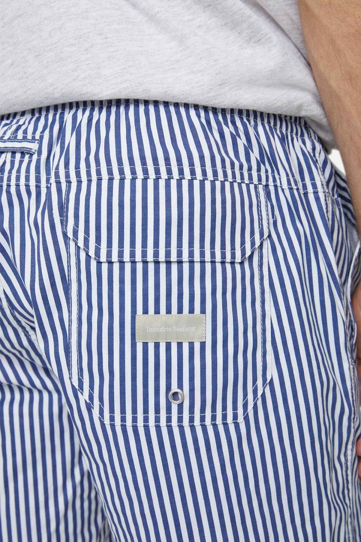 Close-up of someone wearing Industrie The Princeton Bahama Short, showcasing its blue and white stripes and handy back pocket. The tag indicating the comfortable fabric blend suggests premium quality. Their hand rests by their side, accompanied by a light-colored shirt visible at the top.