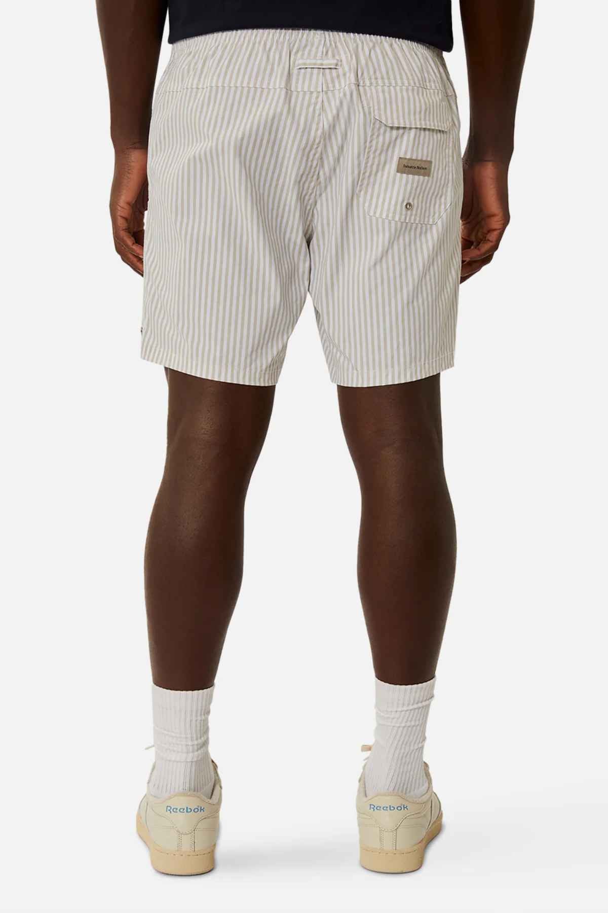 A person dressed in Industrie The Princeton Bahama Shorts, which have cream-colored vertical stripes and a back pocket adorned with a small Industrie branding tag, stands against a plain white background. They pair the shorts with white socks and Reebok sneakers.