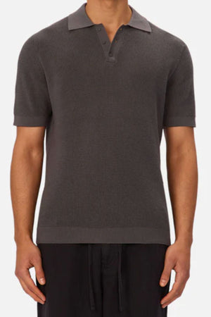 A person is wearing the Industrie The Ravenna Polo—short-sleeve, dark gray, 100% cotton with a partial button placket and collar—paired with black pants, standing against a plain background.
