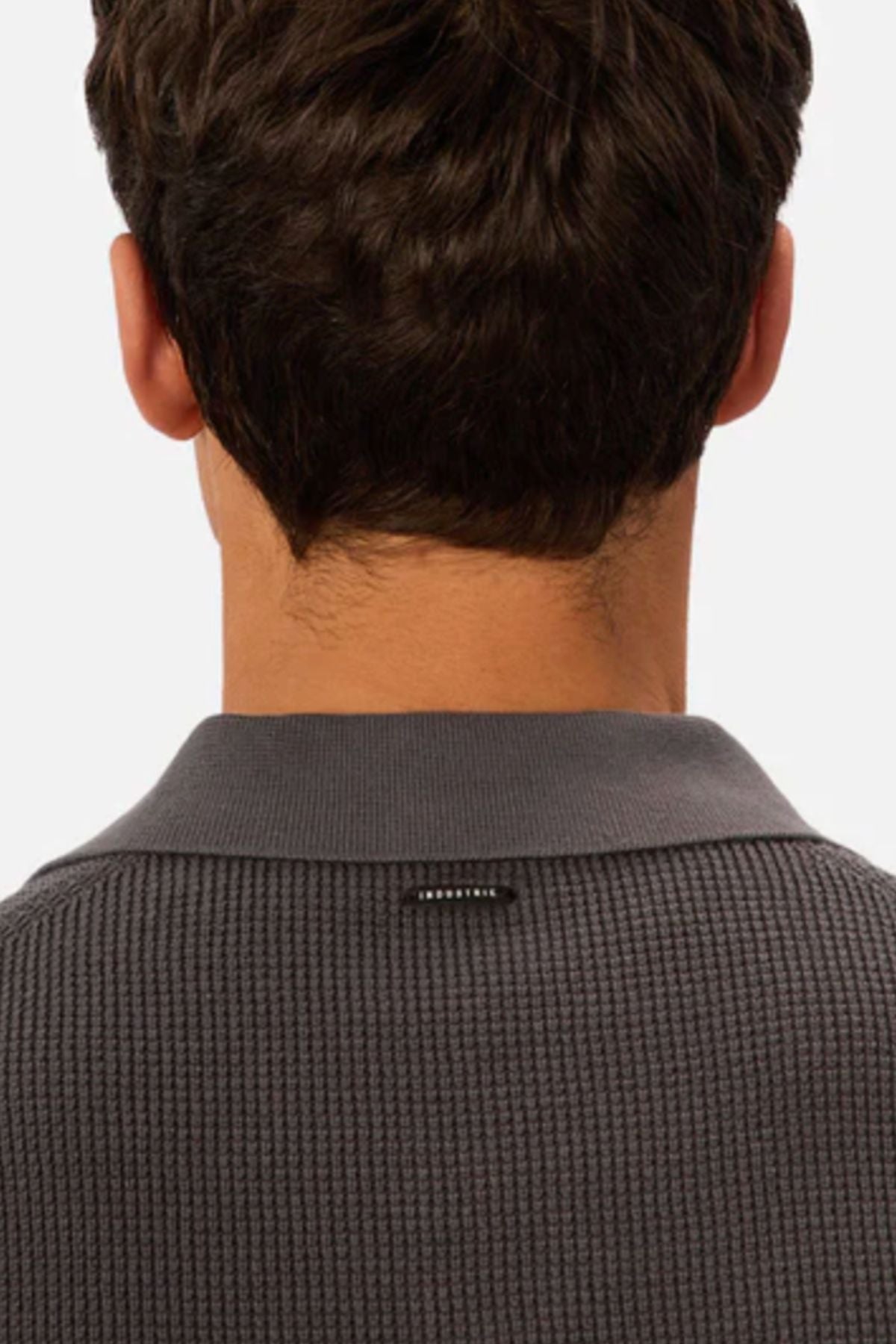 A person with short brown hair is shown from behind, wearing the Industrie The Ravenna Polo in textured dark gray with a small black tag on the back collar. The background is plain and light-colored.