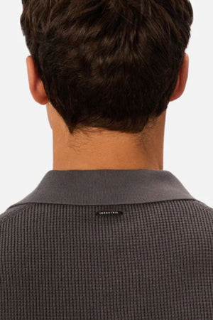 A person with short brown hair is shown from behind, wearing the Industrie The Ravenna Polo in textured dark gray with a small black tag on the back collar. The background is plain and light-colored.