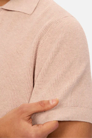 A close-up of a hand gently pinching the sleeve of the Industrie The Ravenna Polo in light pink, showcasing its textured waffle knit fabric.