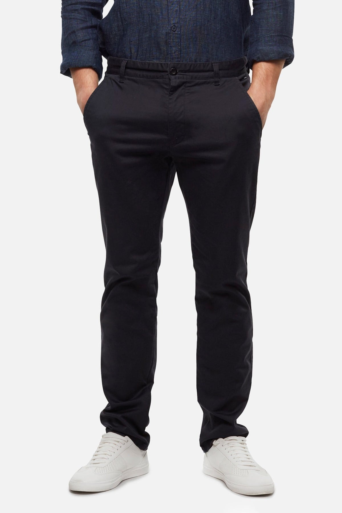 A person stands against a plain light background, wearing a blue long-sleeve shirt, white sneakers, and Industrie The Regular Cuba Chino Pant—a stretch cotton blend chino with a wider leg fit—with hands in pockets.