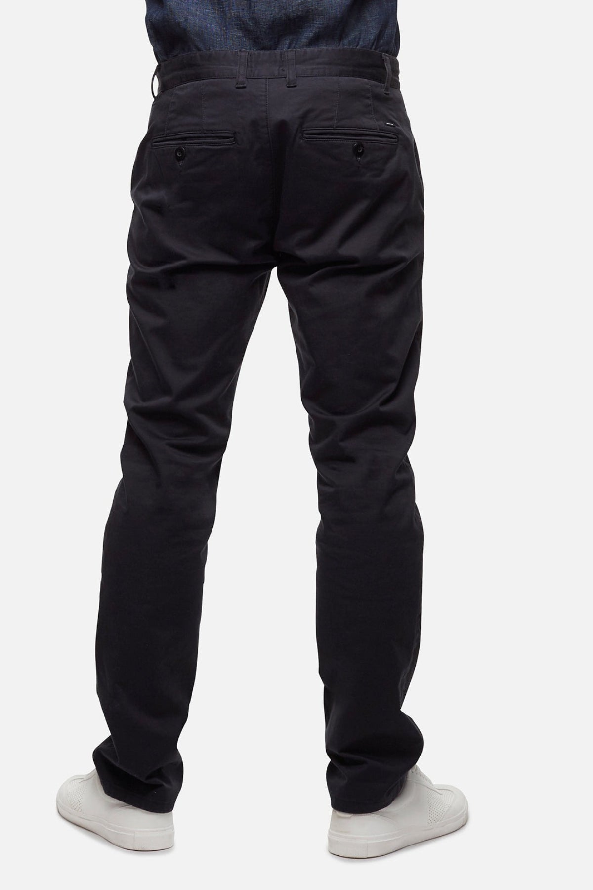 A person stands facing away, hands in pockets, wearing Industrie The Regular Cuba Chino Pant in a stretch cotton blend with a wider leg fit and white sneakers against a plain, light-colored background.