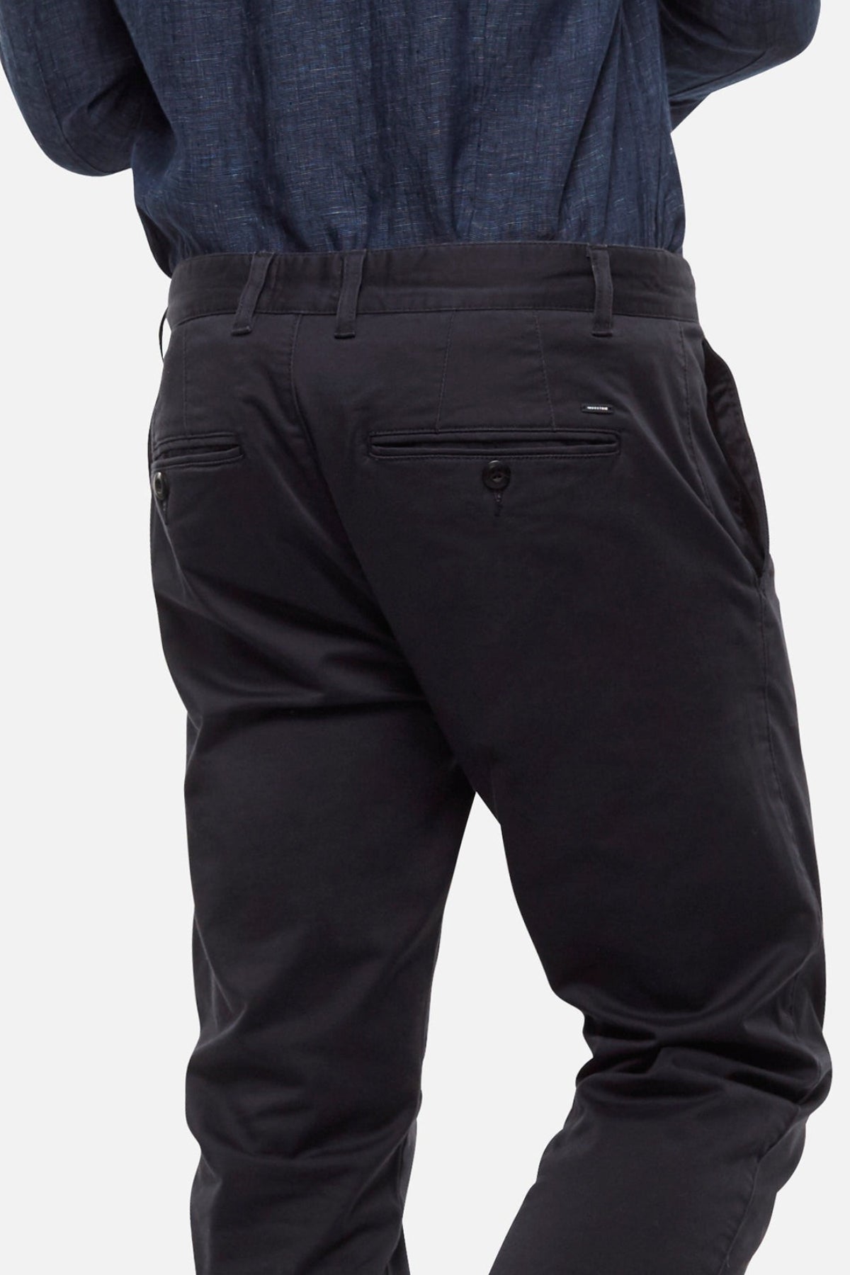 A person is shown from the back wearing Industrie The Regular Cuba Chino Pant in a dark color with a wider leg fit and a blue shirt, hands partially visible and legs slightly bent, set against a plain light background.