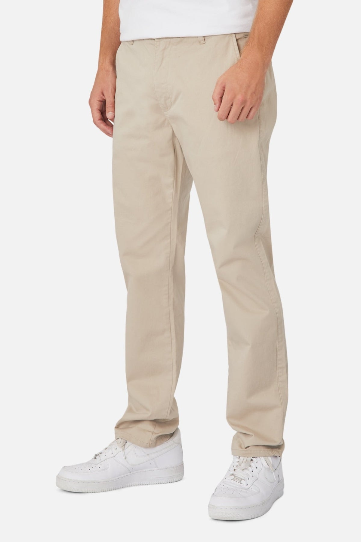 A person is standing against a plain background wearing the Industrie The Regular Cuba Chino Pant in beige and white sneakers. The image captures them from the upper thigh downward, with their hands relaxed at their sides, showcasing the wider leg fit and stretch cotton blend fabric of the chinos.