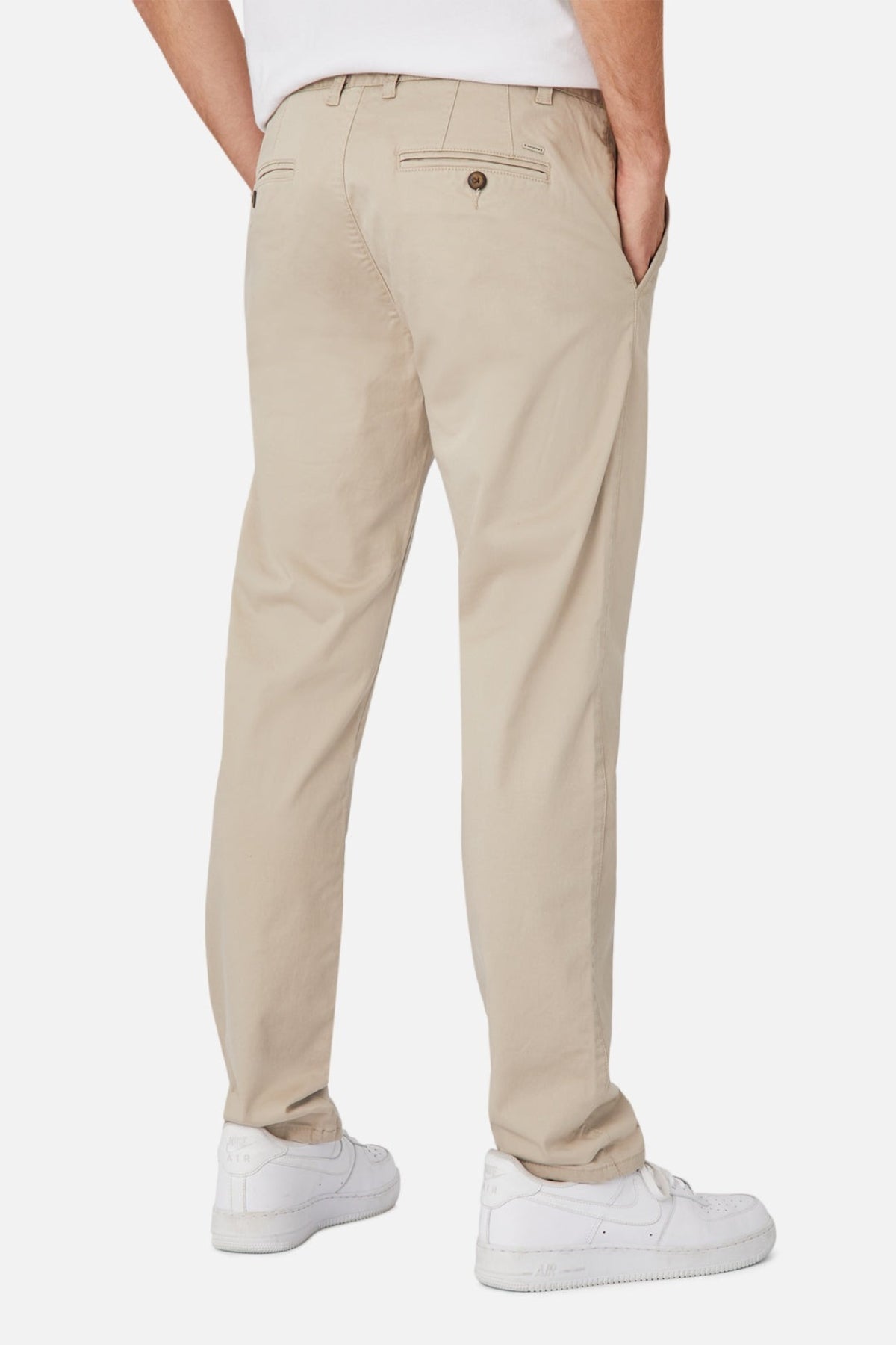 A person stands with hands in pockets, sporting beige Industrie The Regular Cuba Chino Pant made from a stretch cotton blend and white sneakers. The chino pants, known for their wider leg fit, feature back pockets with button closures. The background is white.
