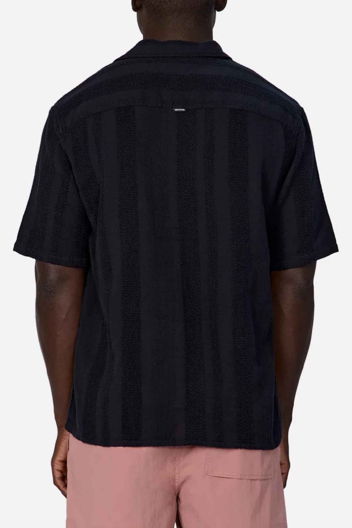 From the back view, the individual is wearing an Industrie The Salamanca Short Sleeve Shirt in a black short-sleeve style featuring subtle vertical stripes. Paired with light pink shorts, this cotton-linen blend ensemble stands out against a plain white background, making it ideal for enhancing any summer wardrobe.