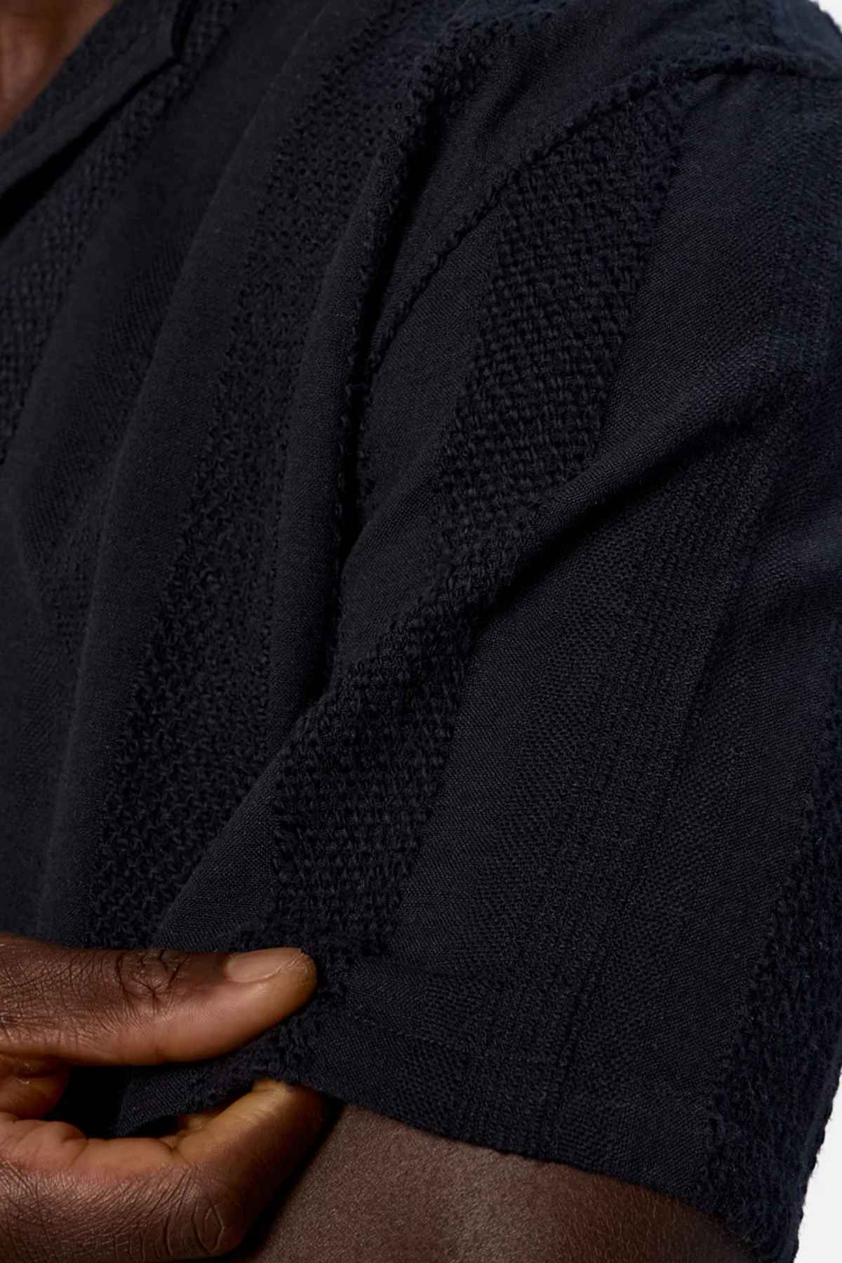Close-up of a person wearing the Industrie The Salamanca Short Sleeve Shirt, crafted from a textured cotton-linen blend. Their hand gently pulls on the cuff of the short sleeve, highlighting its subtle vertical patterns. Ideal for your summer wardrobe, set against a plain white background.