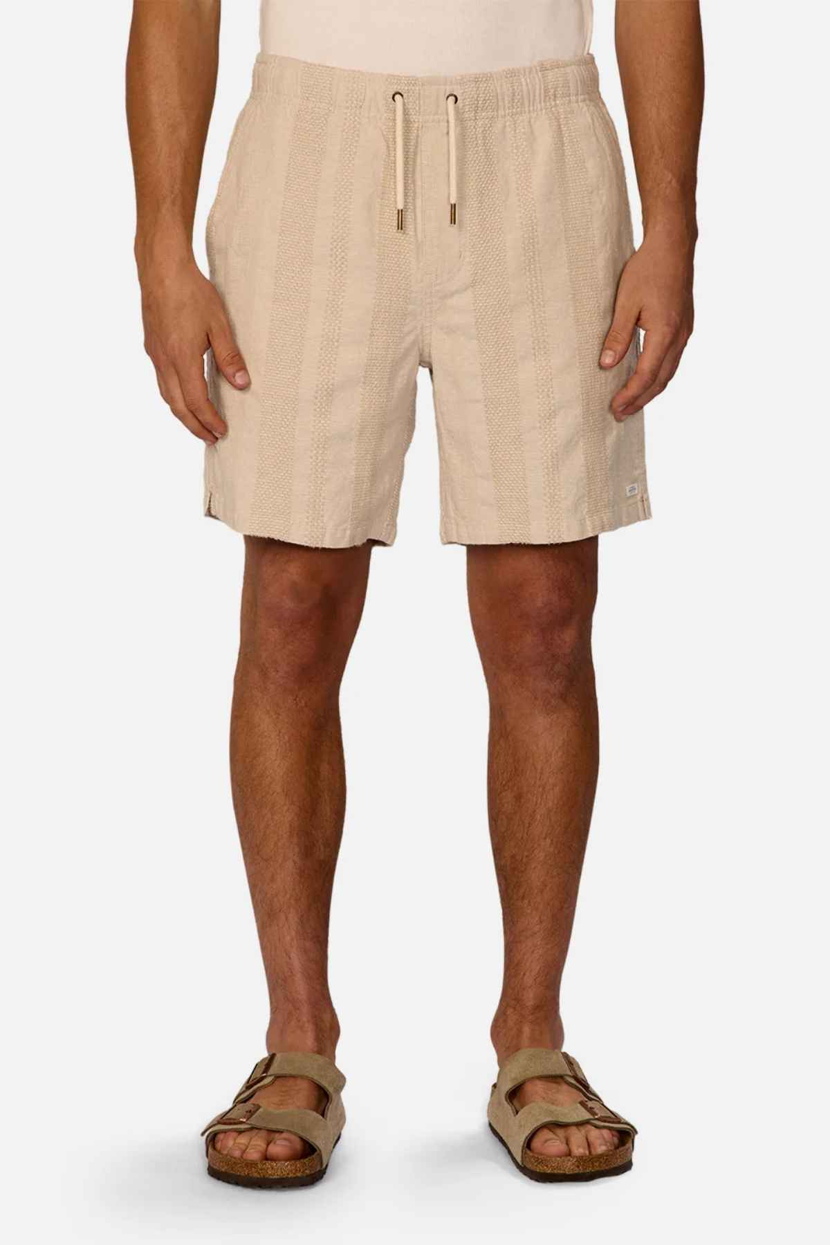 A person wearing light beige Industrie The Salamanca Shorts, featuring a tailored fit and vertical texture lines, stands against a plain background. Made from a cotton linen blend, these shorts are perfectly paired with brown sandals.