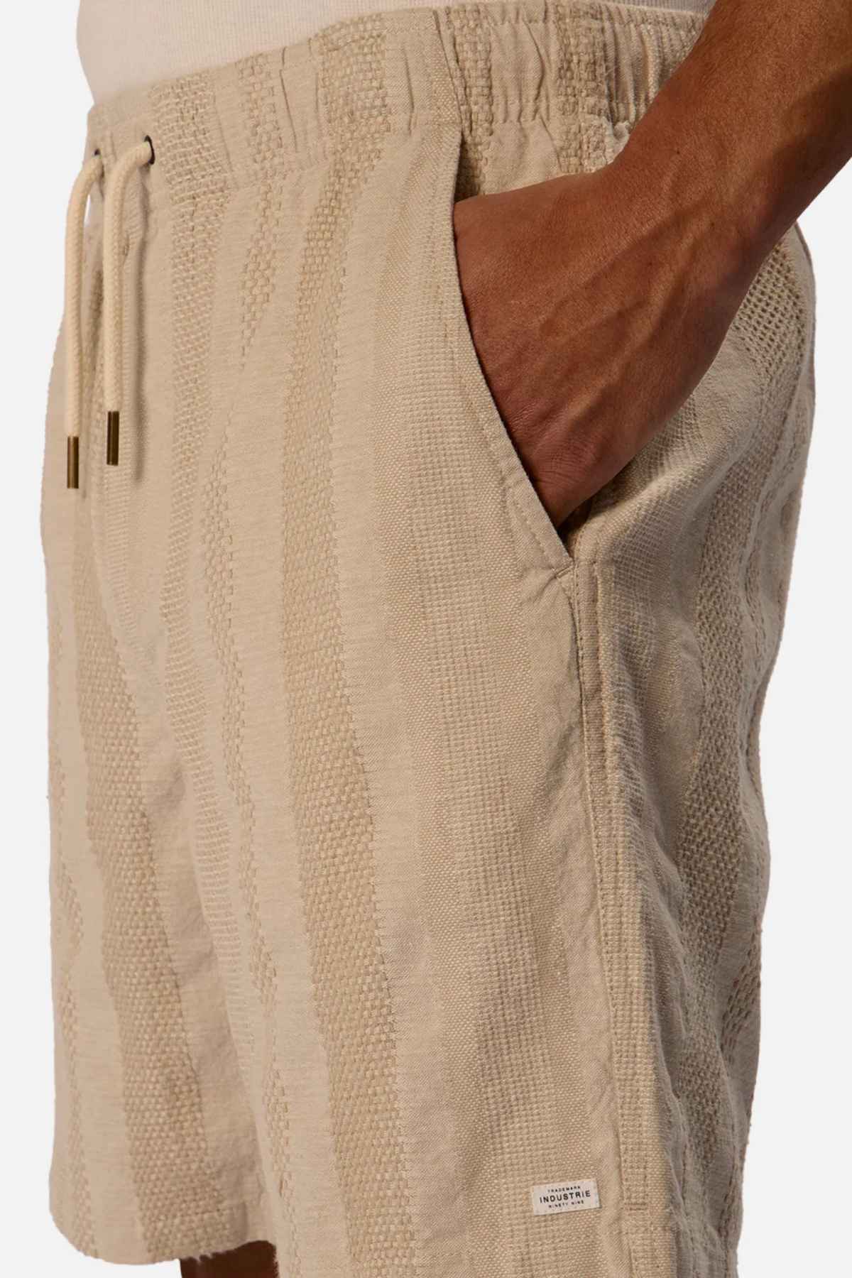 A close-up image focuses on someone wearing the Industrie The Salamanca Short in beige, highlighting its tailored fit and textured cotton linen blend, with a relaxed hand in the pocket. The shorts display a subtle vertical pattern and have a small label near the hem, all set against a plain white background.