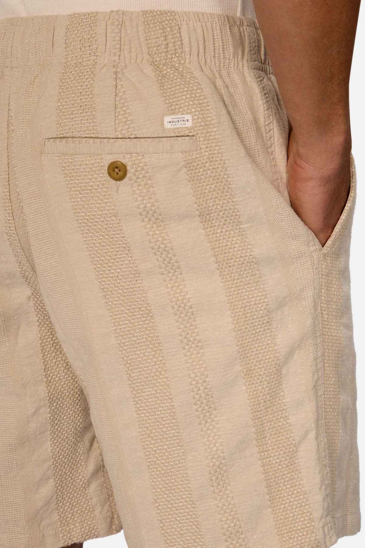 A close-up of a person wearing Industrie The Salamanca Short in beige with vertical stripes. The cotton-linen blend design features an elastic waistband, a brown button on the back pocket, and a white label above it. The tailored fit perfectly complements the hand resting in the side pocket.