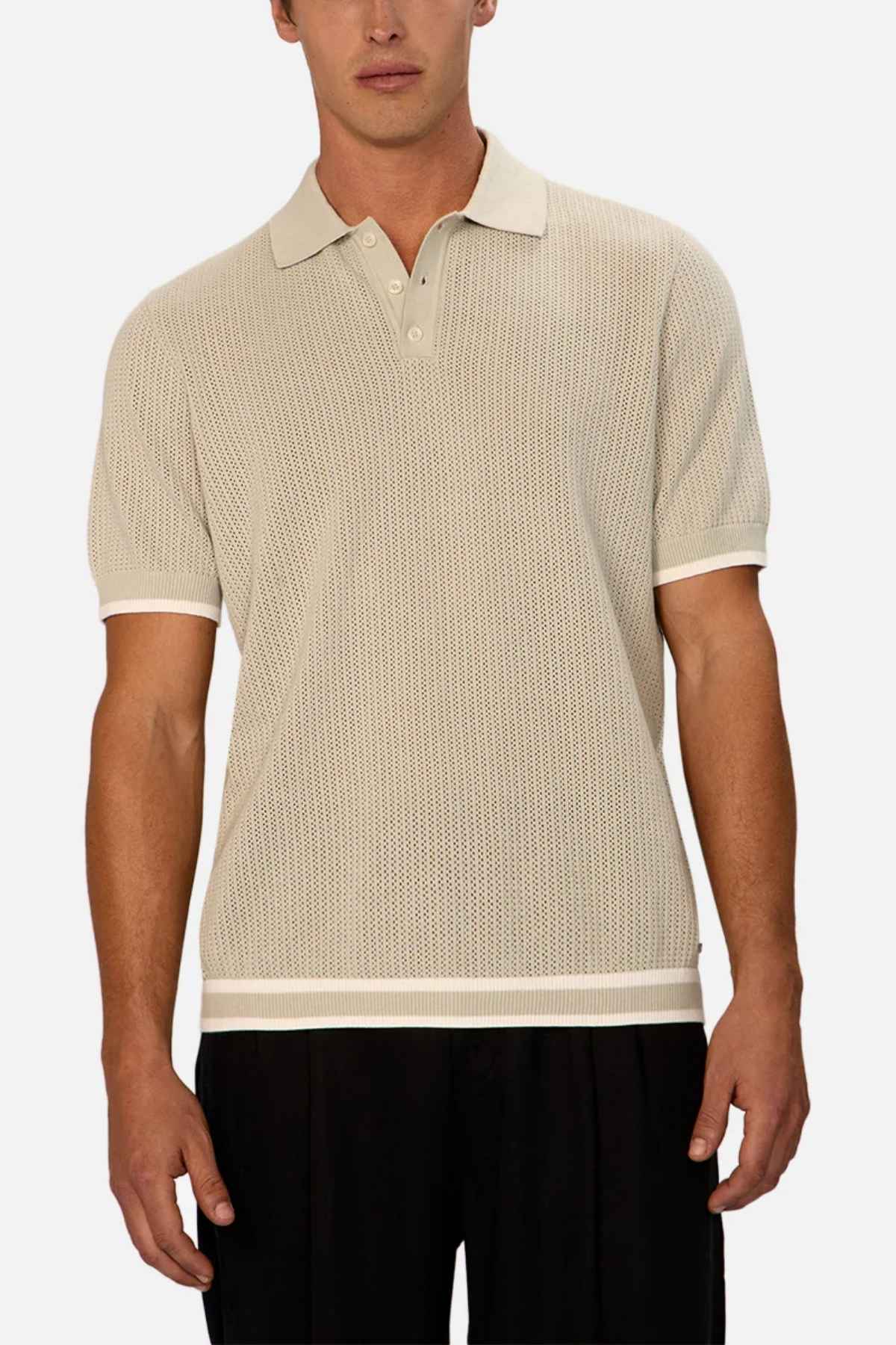 A person is dressed in a beige Industrie The Serrano Polo, crafted entirely from cotton and showcasing a cable knit design along with short sleeves. It's paired with black pants, and the shirt's textured pattern is highlighted against the plain white backdrop.