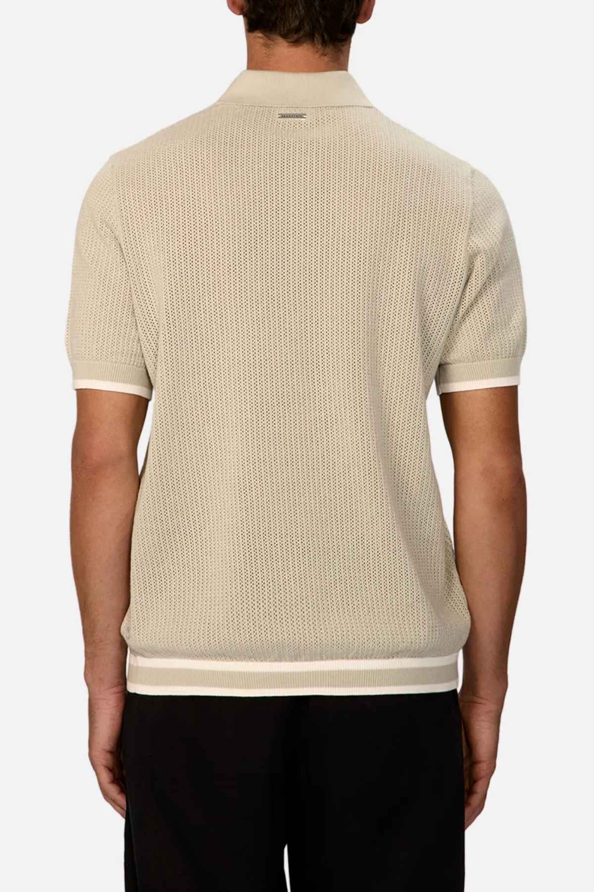 The image depicts a person from behind wearing the Industrie The Serrano Polo, a light beige short-sleeved polo shirt made from 100% cotton. It is perfectly complemented by black pants. The shirt showcases a textured pattern with ribbed cuffs and a classic collar, providing an effortlessly stylish appearance.