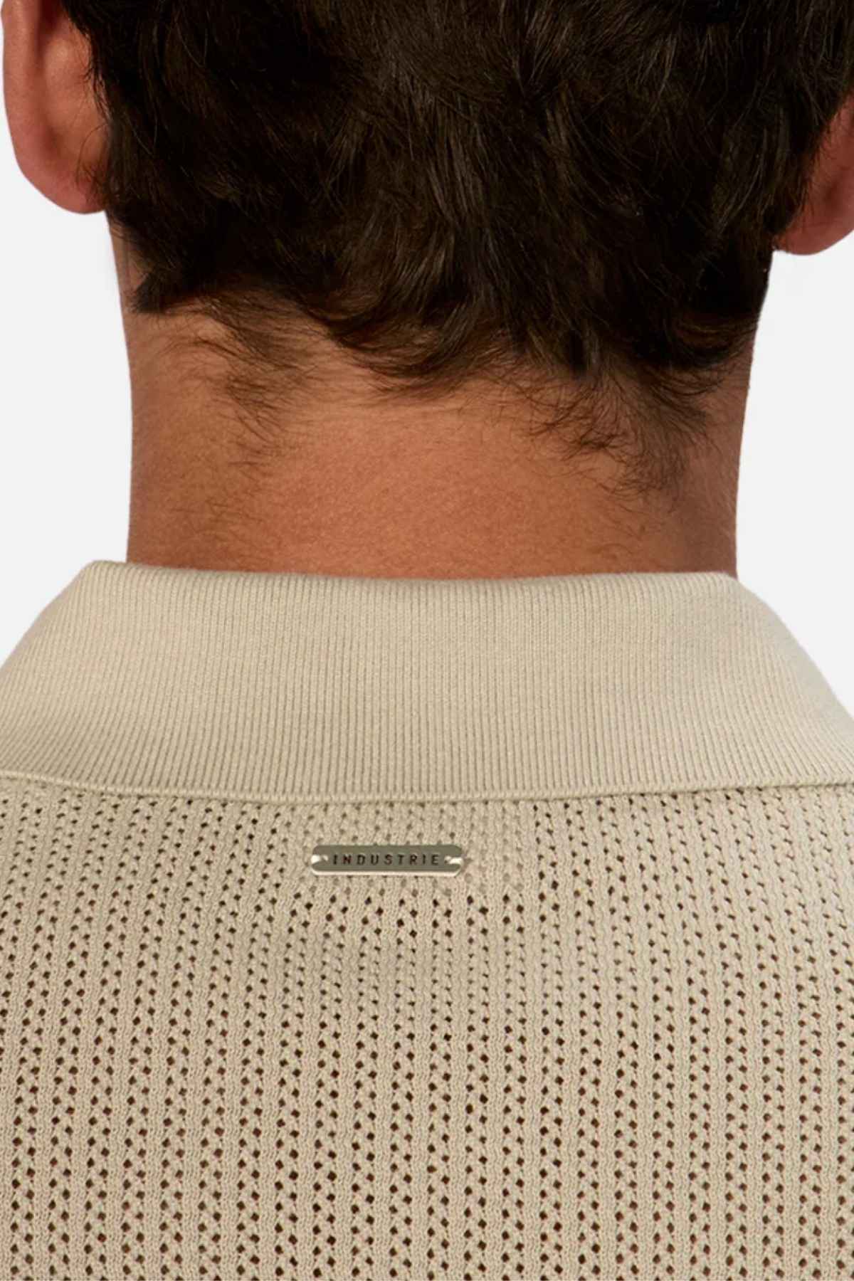 Close-up of the back of a person's head and neck, adorned with the Industrie The Serrano Polo in beige, featuring a perforated design. Made from 100% cotton, the collar tag is emblazoned with "INDUSTRIE." The individual has short, dark hair.