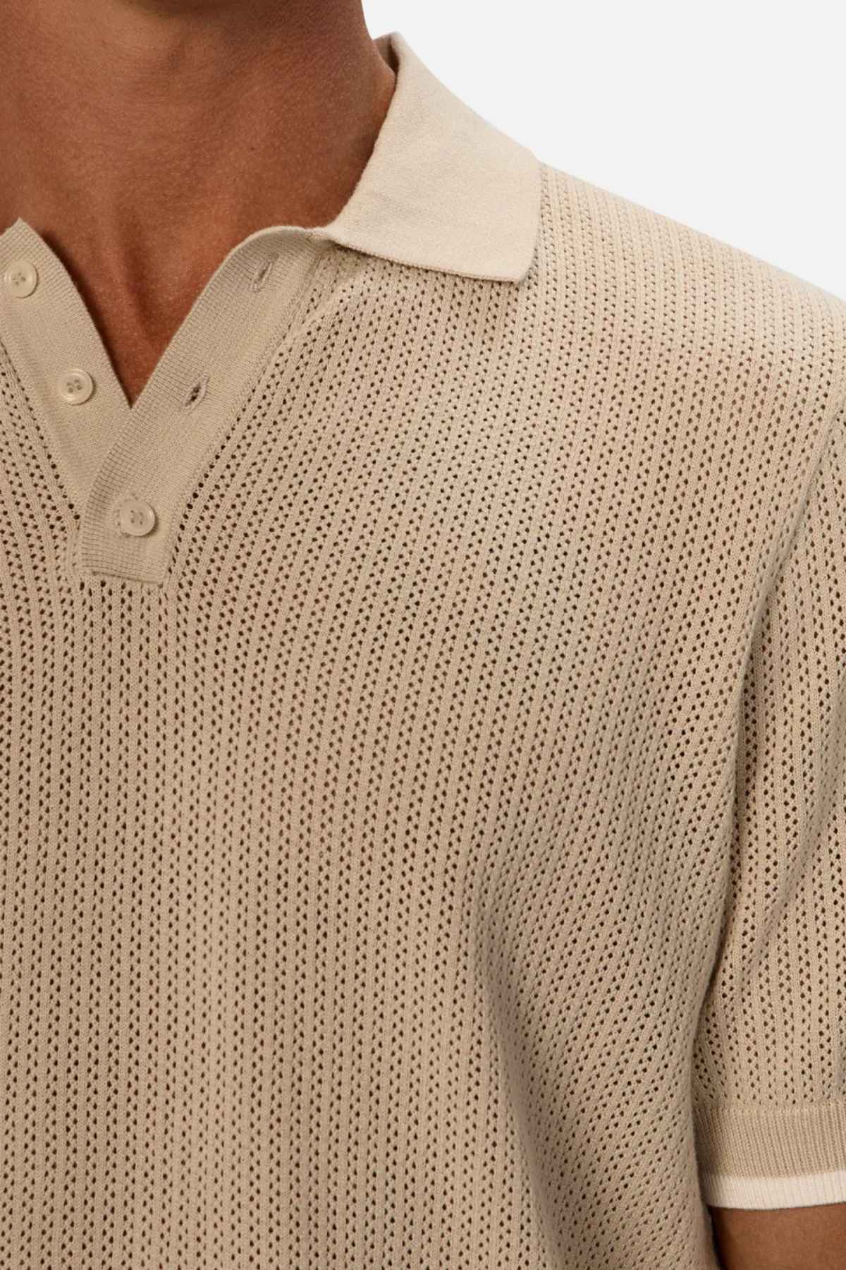 A close-up of someone wearing the Industrie The Serrano Polo showcases a sophisticated beige knit design with a collar and three buttons. The textured fabric is enhanced by a perforated pattern and subtle leather trims, adding an elegant touch.