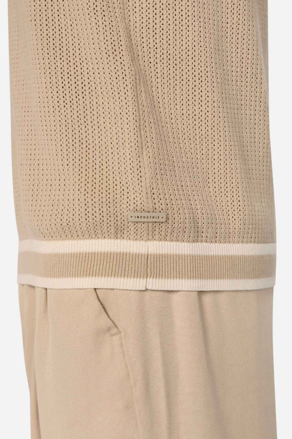 Close-up of a beige cable knit garment with a perforated design, featuring white and beige trim along the lower edge adorned with the word "INDUSTRIE" in small letters. Below, matching beige pants enhance the chic look of the Industrie The Serrano Polo ensemble.