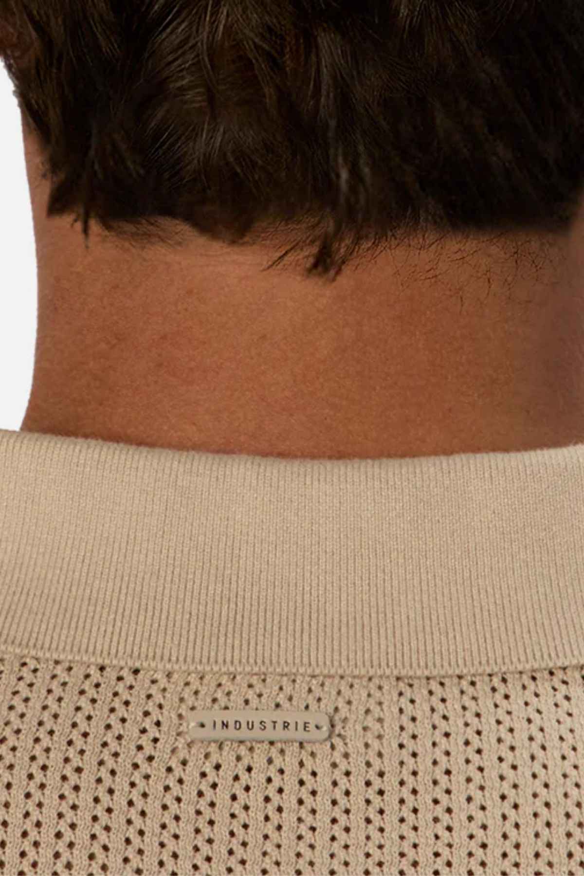 Close-up of the back of a person's neck and shoulders wearing a beige polo. The Industrie The Serrano Polo features a small rectangular label with the word "INDUSTRIE" sewn near the top, adding a touch of classic style. The fabric showcases a textured, knitted pattern.