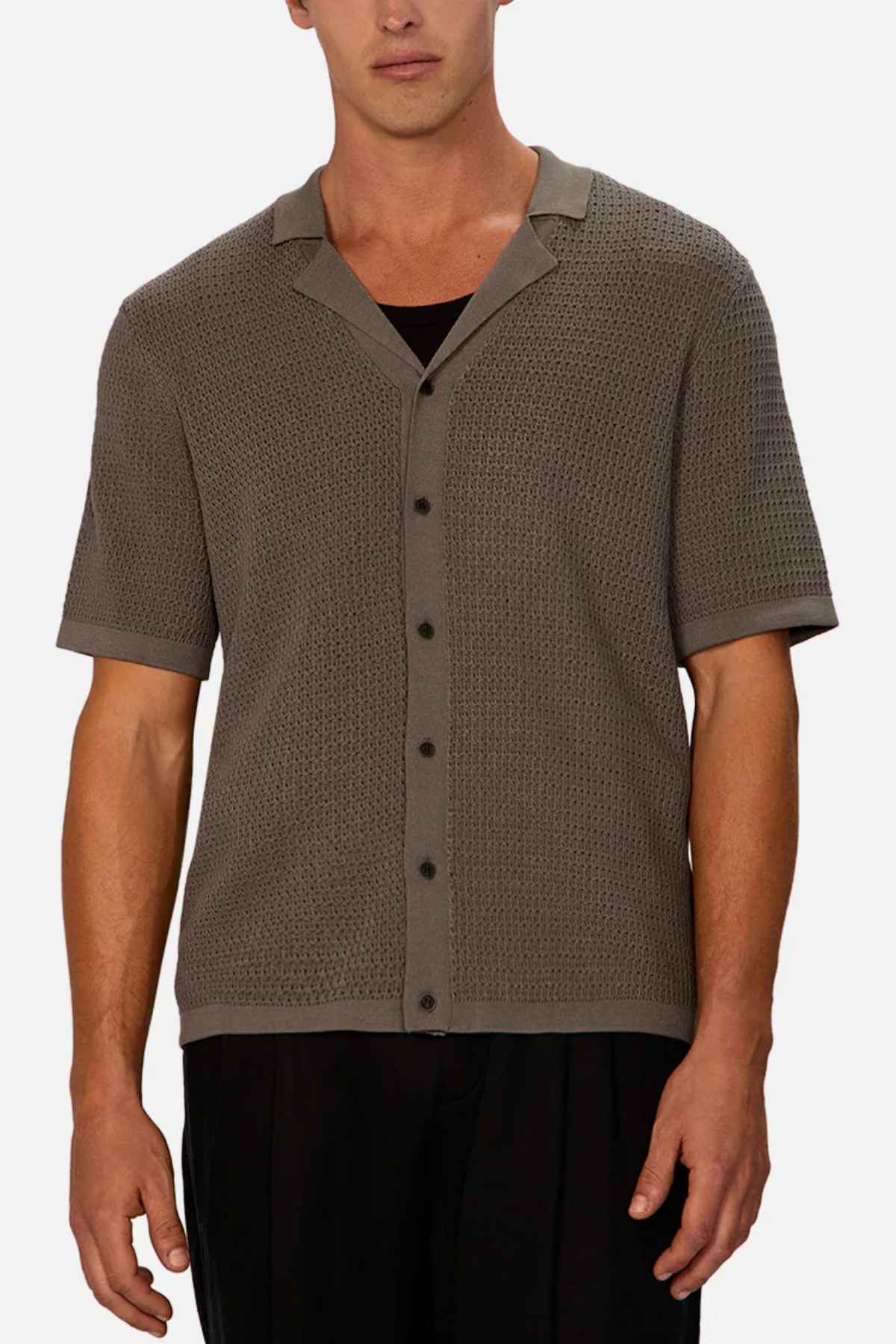 A man wears the Industrie The Sevona Short Sleeve Shirt, featuring a textured, short-sleeve, button-up design in a neutral grey hue over a black undershirt. Offering a relaxed fit perfect for any summer wardrobe, he pairs it with dark pants against a plain white background.