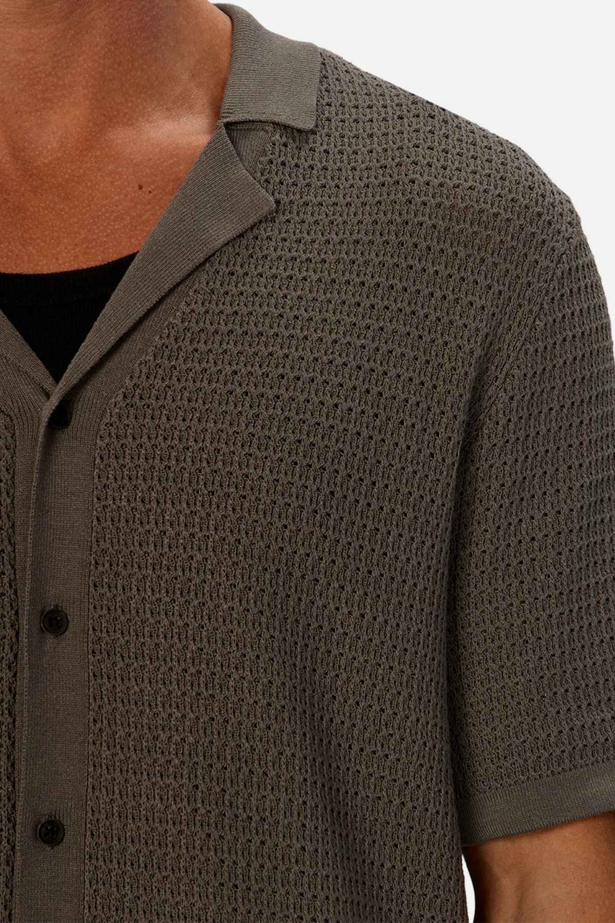 Close-up of a person wearing the Industrie The Sevona Short Sleeve Shirt, a textured, knitted olive green short-sleeve button-up with a relaxed fit over a black undershirt. Perfect for any summer wardrobe, it features a woven pattern and spread collar with black buttons, focusing on the upper torso area.